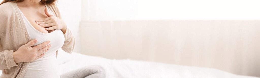 woman sitting on bed massaging right breast wearing white tank top and beige sweater